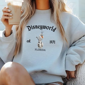 Puede incluir: Una sudadera de color azul claro con un gr&aacute;fico negro de Olaf de Frozen. El gr&aacute;fico incluye el texto "Disneyworld est. 1971 Florida".