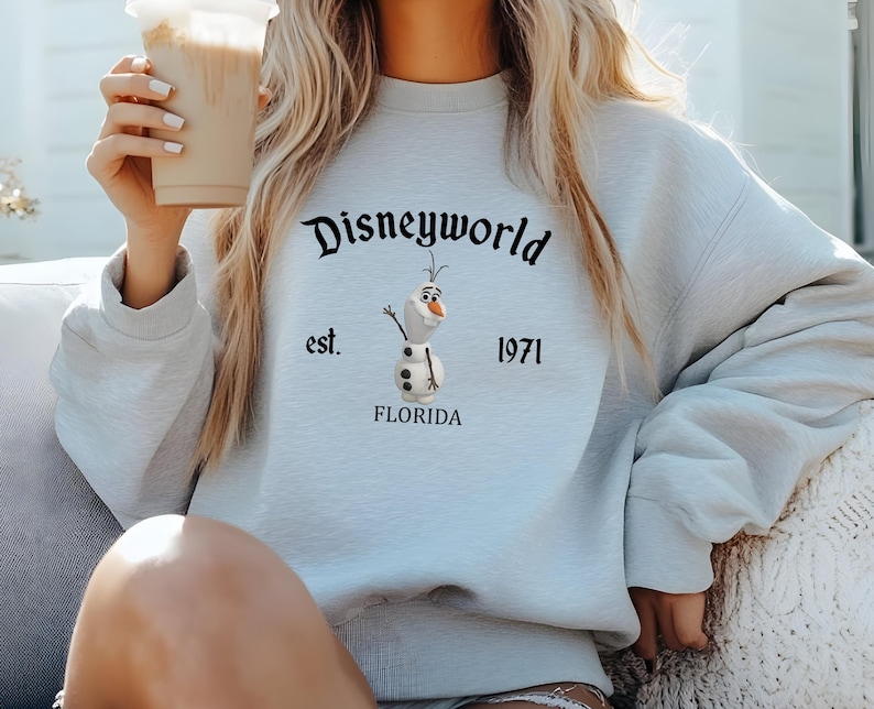 Puede incluir: Una sudadera de color azul claro con un gr&aacute;fico negro de Olaf de Frozen. El gr&aacute;fico incluye el texto "Disneyworld est. 1971 Florida".