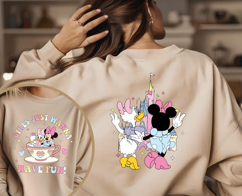 May include: A light beige sweatshirt with a graphic print of Minnie Mouse and Daisy Duck in front of a pink castle. The text "Girls Just Wanna Have Fun!" is printed on the front of the sweatshirt.