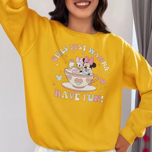 May include: Yellow sweatshirt with a graphic print of Mickey Mouse and Minnie Mouse in a teacup. The text on the sweatshirt reads "Girls Just Wanna Have Fun!"