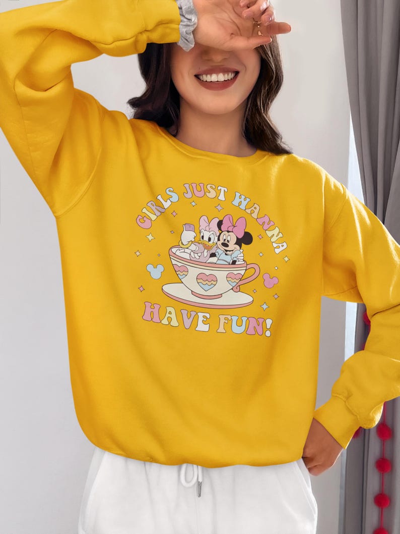 May include: Yellow sweatshirt with a graphic print of Mickey Mouse and Minnie Mouse in a teacup. The text on the sweatshirt reads "Girls Just Wanna Have Fun!"