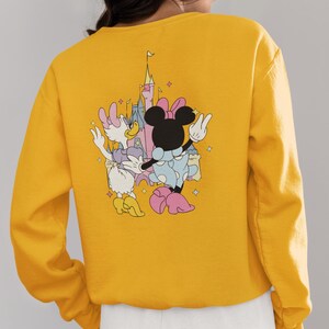 May include: Yellow sweatshirt with a graphic print of Minnie Mouse and Daisy Duck in front of a pink castle. The characters are wearing pink and blue outfits and are holding hands.