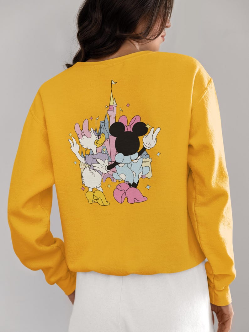 May include: Yellow sweatshirt with a graphic print of Minnie Mouse and Daisy Duck in front of a pink castle. The characters are wearing pink and blue outfits and are holding hands.