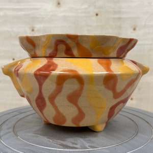 May include: A ceramic planter with a wavy, abstract design in shades of brown, yellow, and orange. The planter has a wide, round base and a smaller, round top.