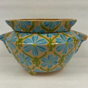 May include: A ceramic planter with a matching lid. The planter is beige with a repeating pattern of blue flowers and green vines. The planter has three small feet and two handles. The lid has a similar floral design.