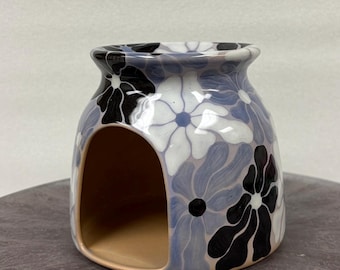 May include: A ceramic wax warmer featuring a rounded shape with a wide opening at the front. The exterior is decorated with a repeating floral pattern in shades of white, light blue, and black against a glossy, smooth surface finish.