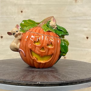 May include: A ceramic pumpkin-shaped candy dish with a jack-o'-lantern face and two small mice peeking out from the top. The pumpkin is orange with a yellow face and green leaves.