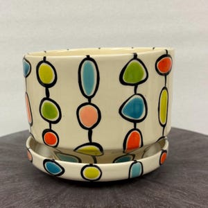 May include: A cream-colored ceramic planter with a matching saucer, featuring a repeating pattern of vertical black lines connecting colorful, rounded shapes in shades of lime green, bright orange, light blue, and soft pink.