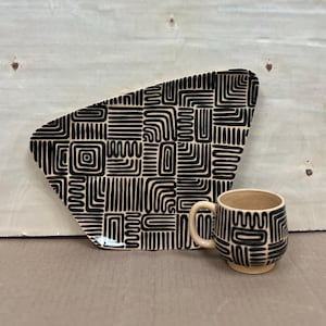 May include: A beige ceramic mug and a matching platter with a black geometric pattern. The mug has a handle and a rounded shape. The platter is an asymmetrical shape.