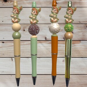May include: Four decorative pens with Peter Pan character toppers. Each pen has a unique design, including a light green, beige, orange, and gold barrel. The toppers feature a Peter Pan figure and decorative beads in various colors and textures.