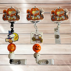 May include: Three retractable badge reels featuring Peanuts Halloween designs. Each reel has a topper with Snoopy and friends near a pumpkin patch, accented with orange pumpkin beads, patterned beads, and charms of Snoopy as a witch or pilot.