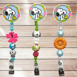 May include: Three Snoopy-themed retractable badge holders. Each features a Snoopy graphic surrounded by flowers. One has a pink flower, another a Snoopy figurine, and the third a sunflower. The badge reels have various colorful beads.