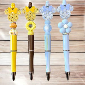 May include: Four decorative pens with unique designs. The pens feature yellow, brown, and blue barrels, topped with various embellishments, including sunflowers, bows, and floral accents. Each pen has a silver tip and decorative elements.