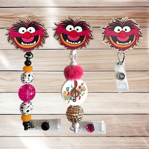 May include: Three badge reels featuring the character Animal from The Muppets with glittery edges. The first has black, orange, music note, and pink beads. The second has a pink pom-pom, a music note disc, and a gold bead. The third has a clear clip.