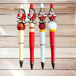 May include: Four decorative pens featuring cartoon dwarf characters. Each pen has a different design, with red, white, and yellow accents. The pens have black ink tips and are set against a light wood grain background.