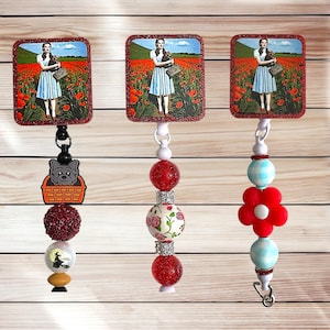 May include: Three retractable badge holders featuring a scene from The Wizard of Oz. Each holder has a rectangular image of Dorothy in a poppy field. The holders have different decorative beads, including a dog in a basket, red glitter beads, and floral designs.