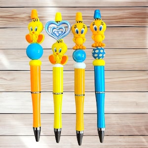 May include: Four decorative pens featuring cartoon character designs. Each pen has a different design, including a yellow bird figure, a blue heart, and a blue and yellow bird figure. The pens have colorful barrels in orange, yellow, and blue.