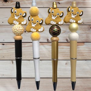 May include: Four decorative pens featuring a Simba character design. Each pen has a different color scheme, including black, white, gold, and beige barrels. The pens are topped with decorative beads and Simba figures.