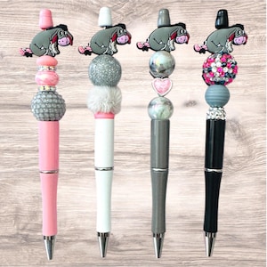 May include: Four decorative pens with unique designs. Each pen features a different color scheme, including pink, white, gray, and black. The pens are topped with a cartoon donkey character and various decorative beads and accents.