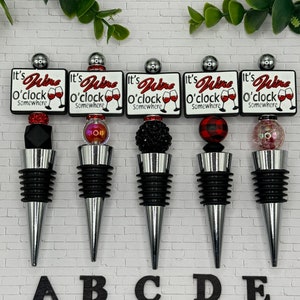 May include: Five silver wine bottle stoppers with a white square design that says "It's Wine O'clock Somewhere". Each stopper has a different embellishment: black beads, red beads, black rhinestones, red and black plaid fabric, and iridescent glitter.