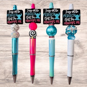 May include: Four decorative pens with black rectangular toppers featuring the text "I may not be perfect but I'm always me" and a cartoon character. The pens have colorful barrels and decorative tops with beads and rhinestones.