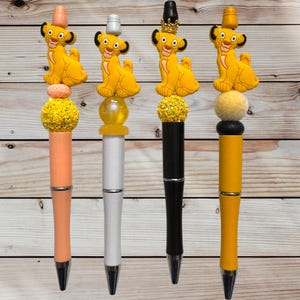 May include: Four decorative pens featuring a yellow lion cub design. The pens have orange, white, black, and yellow barrels, with silver accents. Each pen has a different topper, including a crown and various ball-shaped embellishments. The pens are on a wooden background.