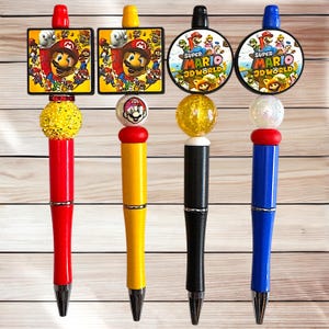 May include: Four colorful pens featuring Super Mario characters. Two pens have square toppers with Mario images, while the other two have round toppers with "Super Mario 3D World" logos. Each pen has a unique color scheme, including red, yellow, black, and blue.
