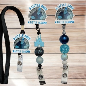 May include: Three badge reels with the text "A LITTLE DARK" and "A LITTLE GLOOMY". One reel has a black lanyard. The other two have decorative beads in shades of blue and gray, with a castle charm.