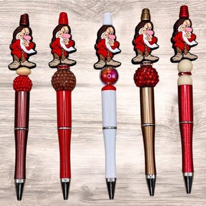 May include: Five decorative pens with character toppers. Each pen features a different design, including red, white, and gold barrels, and various bead accents. The toppers depict a cartoon character in a red coat.