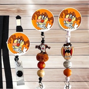 May include: Three badge reels featuring the Tasmanian Devil character. Two reels have circular tops with Taz in a tornado on an orange background. One reel has a black lanyard. The other two feature beaded dangles with Taz charms and orange, brown, and white beads.