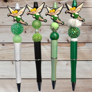 May include: Four decorative pens with green and white accents. Each pen features a different design, including a Tinkerbell figure, glittery beads, and a variety of green shades. The pens have silver and black tips and are set against a wooden background.