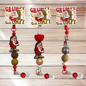 May include: Three retractable badge holders featuring the character Grumpy from Snow White. Each holder has a Grumpy image with the word "Grumpy" in red and gold. The holders have red, white, and brown beads and a red gem.