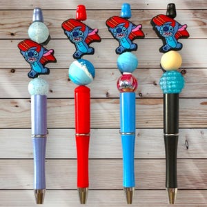 May include: Four decorative pens with Stitch character toppers. The pens have different colored barrels: purple, red, blue, and black. Each pen features a Stitch figure, a decorative bead, and a small topper.