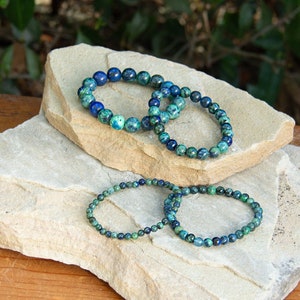 May include: Four beaded bracelets made of blue and green chrysocolla stones. The bracelets are stacked on top of each other on a light brown rock.
