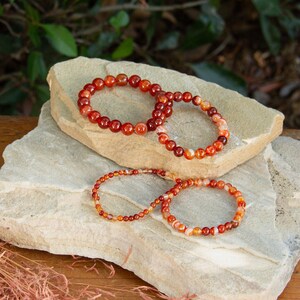 May include: Three carnelian gemstone bracelets on a stack of light brown stones. The bracelets are made of small, round beads in shades of orange, red, and white.