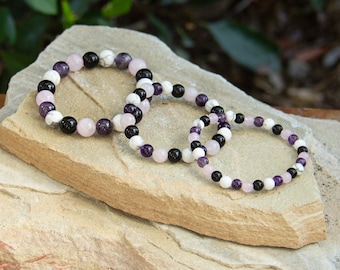 May include: Three beaded bracelets made with amethyst, white howlite, and black onyx stones. The bracelets are stacked on top of each other on a light brown stone surface.