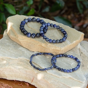 Op de afbeelding: Vier blauwe edelsteen armbanden gestapeld op elkaar op een lichtbruine steen. De armbanden zijn gemaakt van kleine, ronde kralen.