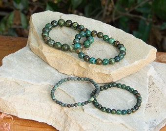 May include: Four green gemstone bracelets stacked on top of each other on a light brown rock surface. The bracelets are made of small, round beads.