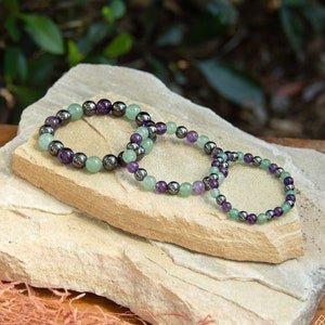 May include: Three bracelets made of amethyst, green aventurine, and hematite beads. The bracelets are stacked on top of each other on a light brown stone surface.