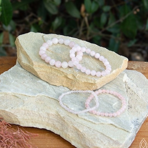 May include: Several rose quartz beaded bracelets are displayed on a light-colored stone surface. The bracelets vary in size and bead diameter, showcasing a soft pink hue. The background features greenery and a wooden surface.