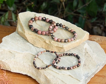 May include: Four bracelets made of pink and black gemstone beads are displayed on two stacked stone slabs. The bracelets are arranged in two pairs, with the larger bracelets on the top slab and the smaller bracelets on the bottom slab.