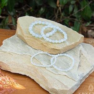 May include: Four white opal beaded bracelets stacked on top of each other on a light brown rock surface. The bracelets are made of small, round beads.
