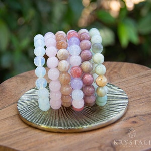 May include: A collection of beaded bracelets in various colors, including white, pink, orange, purple, and green. The bracelets are displayed on a gold-colored, round decorative plate, set on a wooden surface. The bracelets are made of round beads.