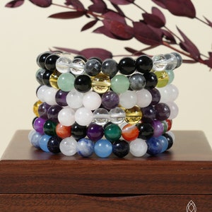 May include: A stack of eight bracelets made with various semi-precious stones, including amethyst, clear quartz, green aventurine, and black onyx. The bracelets are arranged on a wooden box with the text "KRYSTALI CREATIONS" on the front.