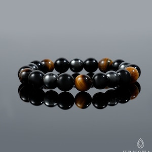 K&ouml;nnte beinhalten: Ein Armband mit schwarzen, braunen und goldenen Perlen. Das Armband besteht aus H&auml;matit-, Tigerauge- und Onyxsteinen.