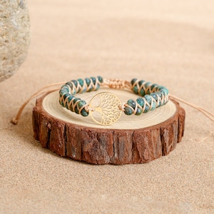 May include: A green and brown beaded bracelet with a gold tree of life charm. The bracelet is on a wooden surface.