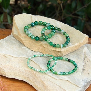 May include: Four green agate beaded bracelets stacked on top of each other on two gray rocks. The bracelets are made of small, round beads.
