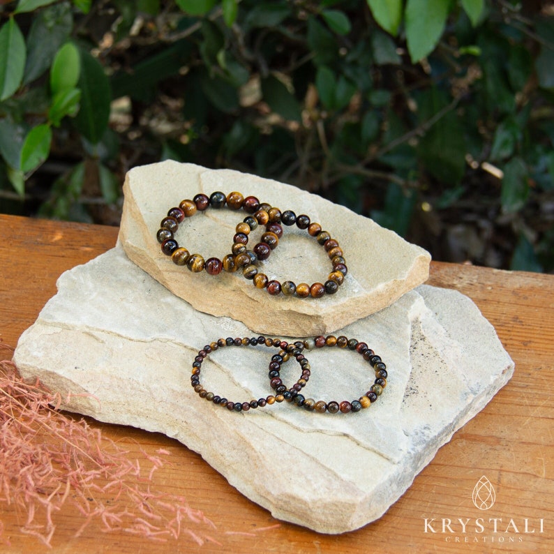 May include: Three tiger's eye gemstone bracelets stacked on top of each other on a light brown stone surface. The bracelets are made of small, round beads in shades of brown, gold, and red.