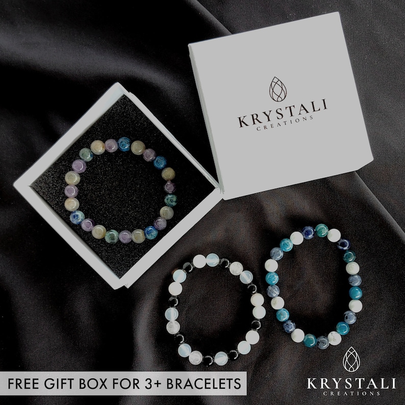 May include: Three beaded bracelets in shades of blue, green, white, and purple are displayed on a black surface. One bracelet is in a white gift box. The text "KRYSTALI CREATIONS" is visible on the box and on the bottom right. The text "FREE GIFT BOX FOR 3+ BRACELETS" is at the bottom.