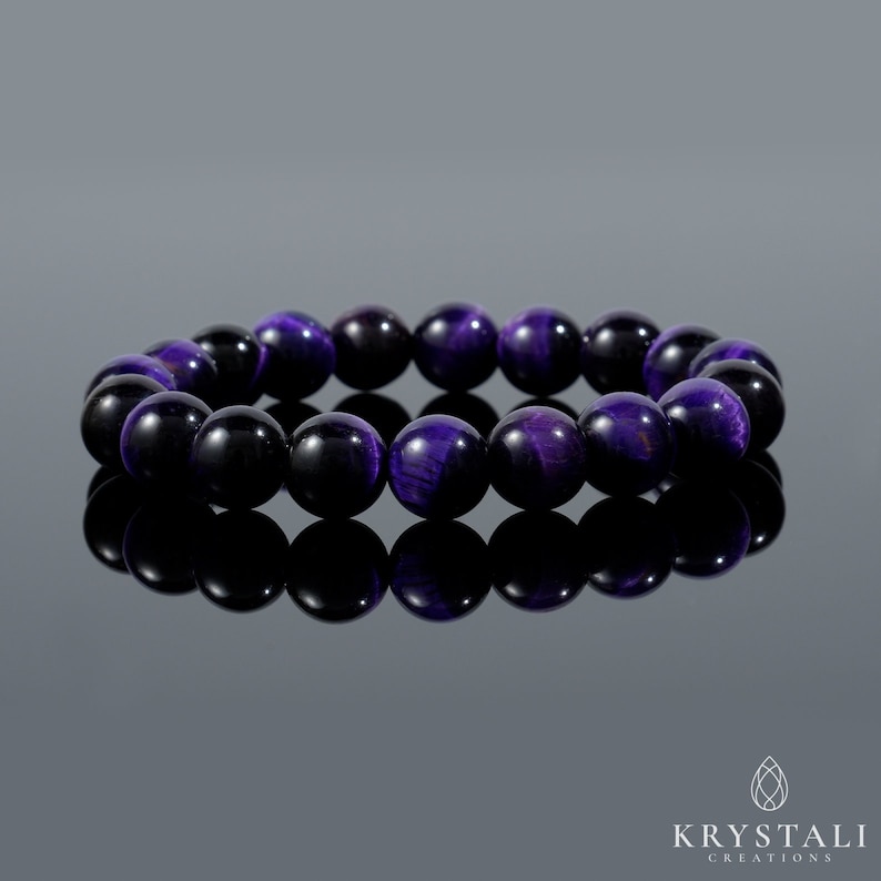 May include: A beaded bracelet featuring polished, round beads in shades of deep purple and black. The bracelet is displayed on a reflective surface, showcasing the jewelry's color and design. The beads appear to be approximately 0.4 inches in diameter.